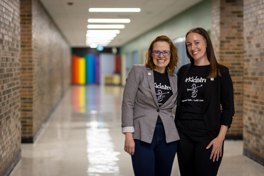 Representing the Co-Leadership of OrKidstra, Ms. Carrie Lauzon, Executive Managing Director (left) and Ms. Danielle Girard, Artistic Managing Director (right) stand together in the hallway of one of OrKidstra's Hubs, located at Ottawa Technical Secondary School in Vanier.
